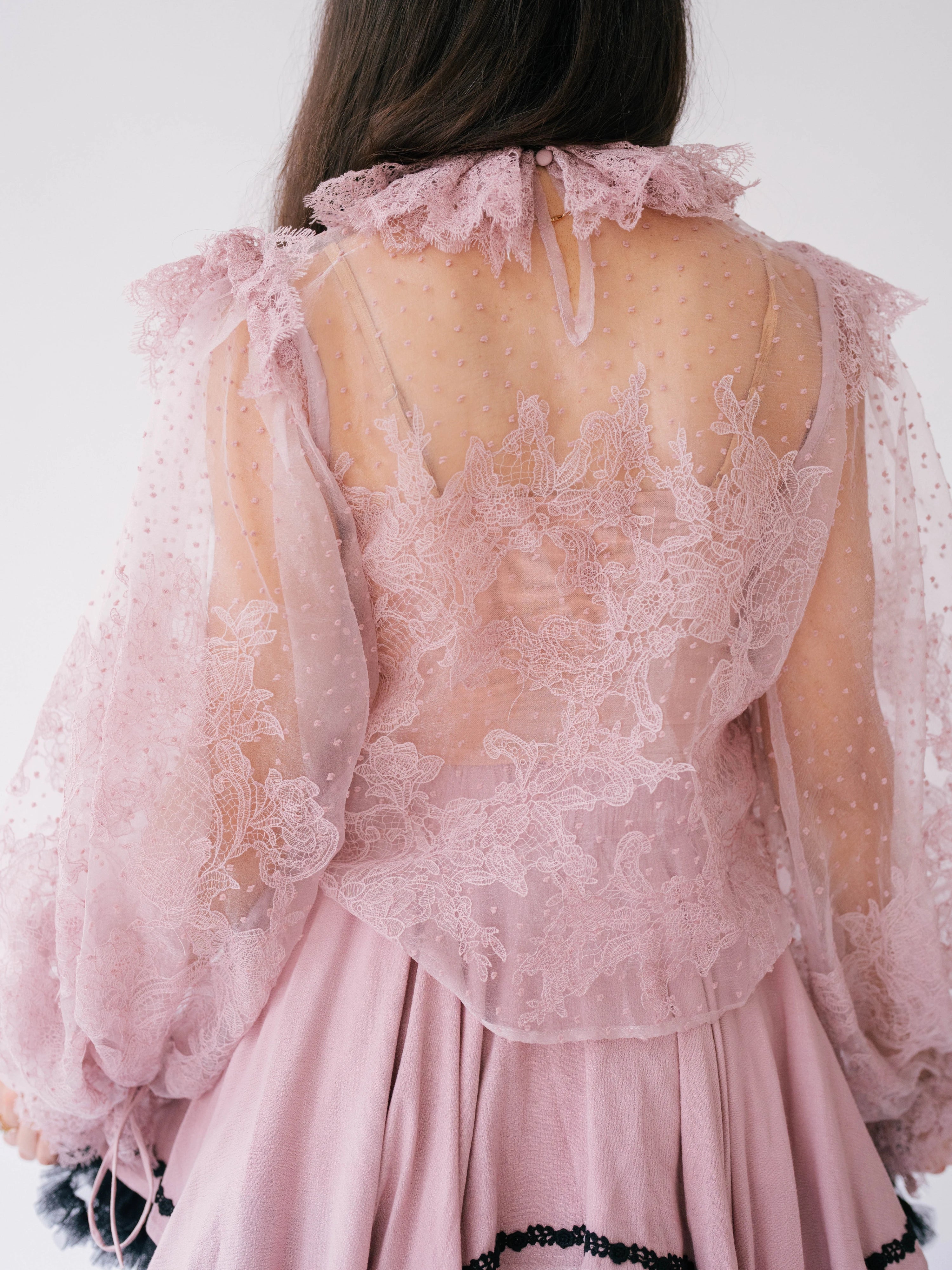 The Eleanor Dusty Pink Lace Blouse is a delicate pink lace blouse with sheer long puff sleeves and is shown close up from the back.