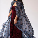 A flowing floral print organza cape worn over a red velvet dress.