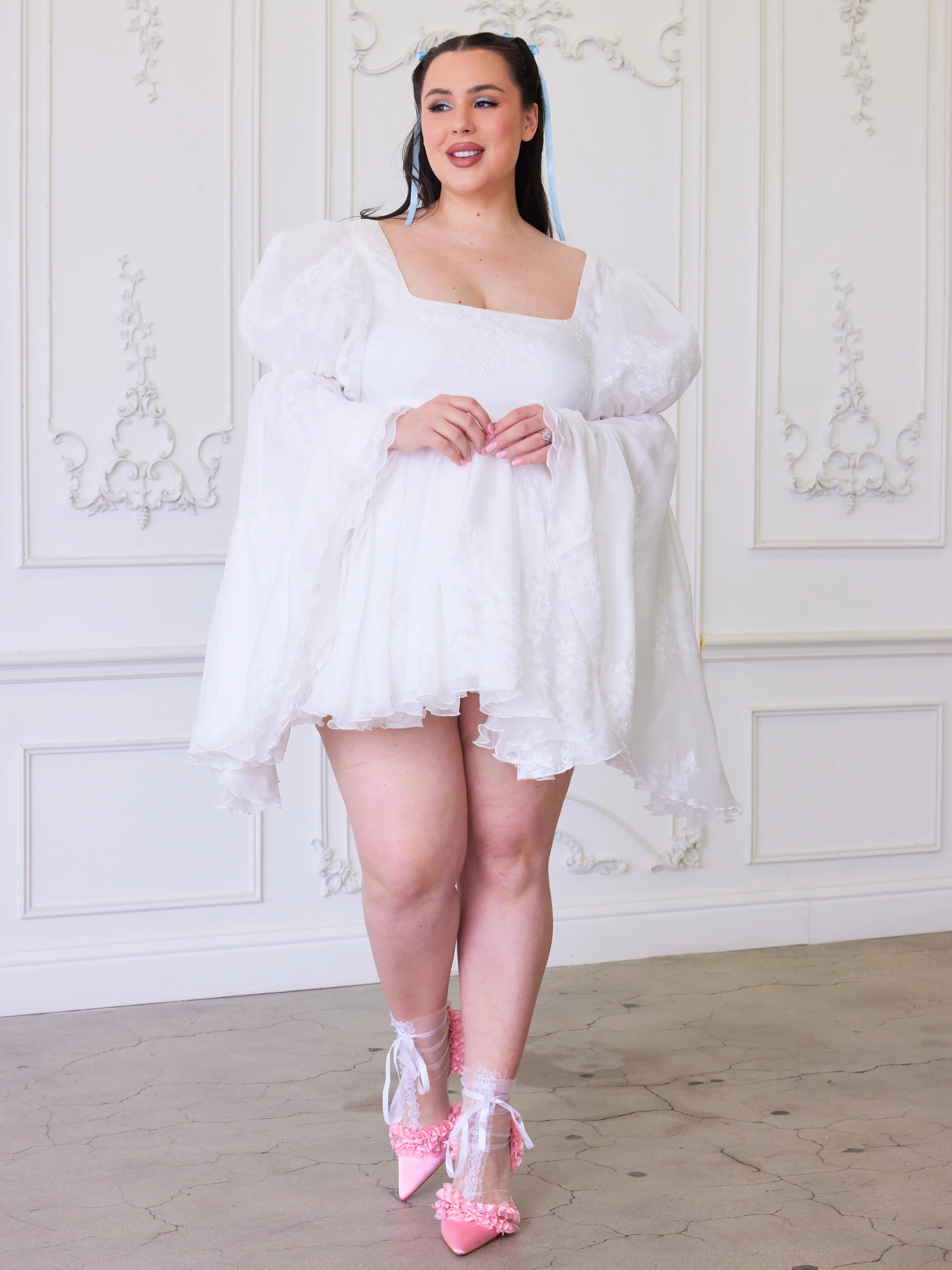 An elegant white mini dress with puffy oversized bell sleeves and pink heels.