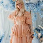 Blonde model poses in the Midnight Symphony Mini Gown surrounded by blue and peach florals, emphasizing its romantic heirloom design.