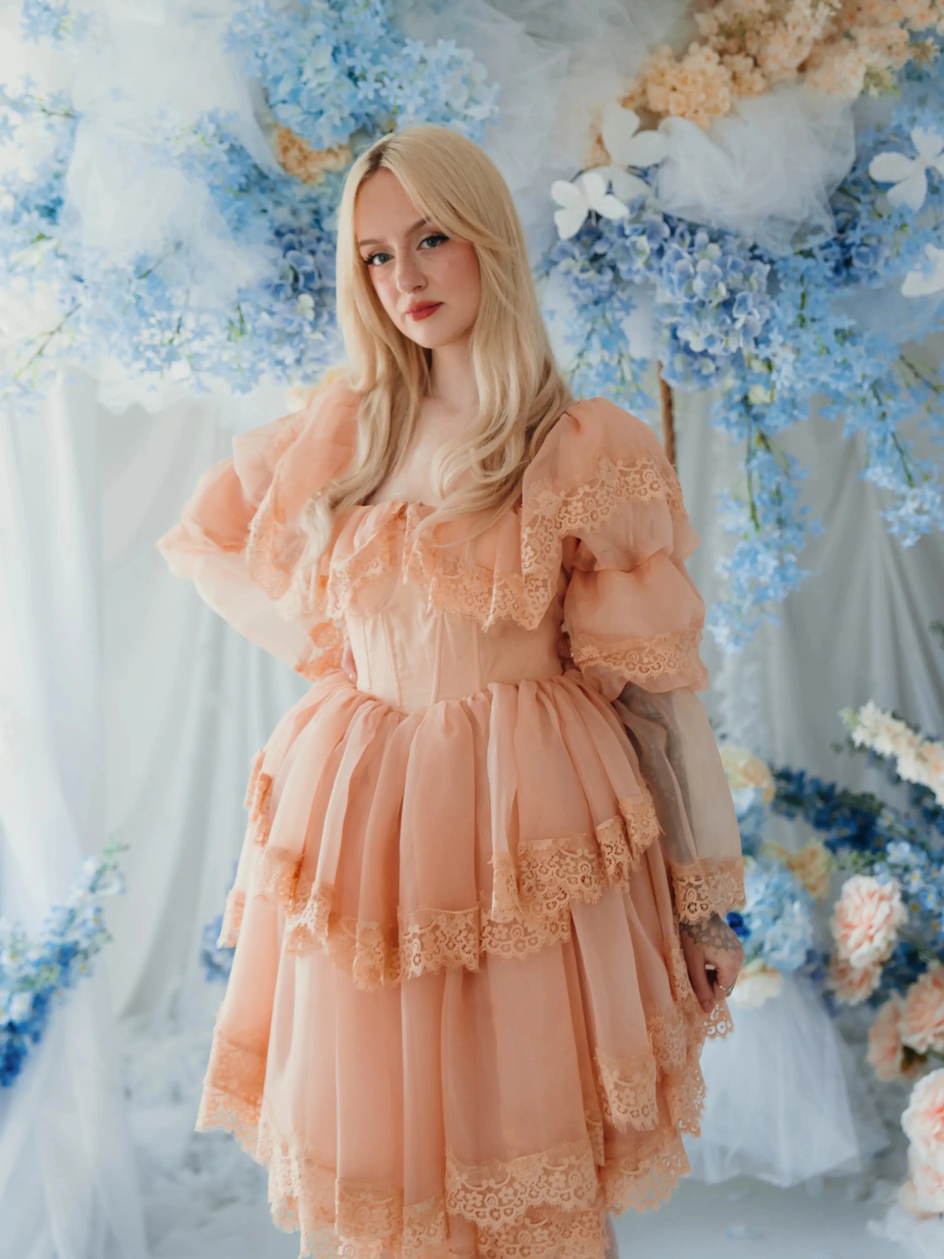 Blonde model poses in the Midnight Symphony Mini Gown surrounded by blue and peach florals, emphasizing its romantic heirloom design.