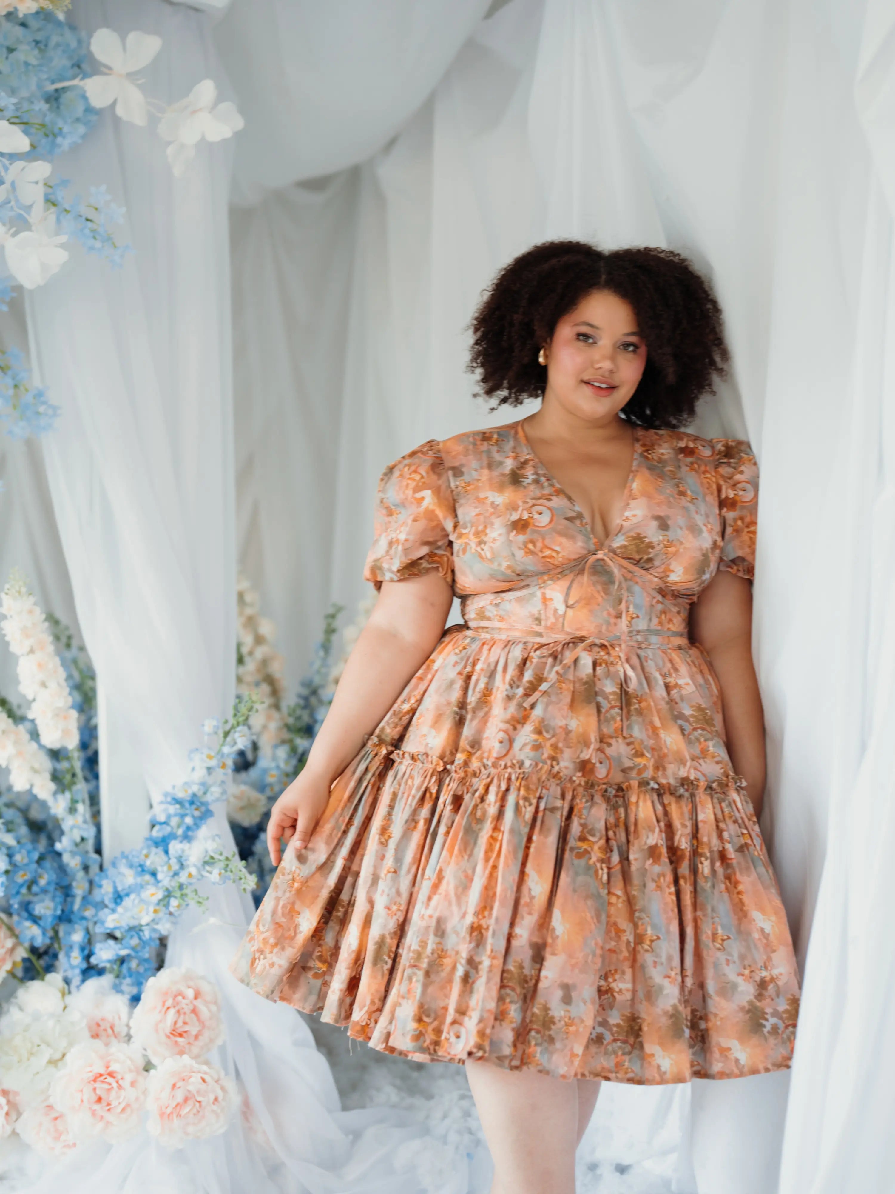 Woman wearing a floral dress in a decorated indoor setting with flowers and white drapes.