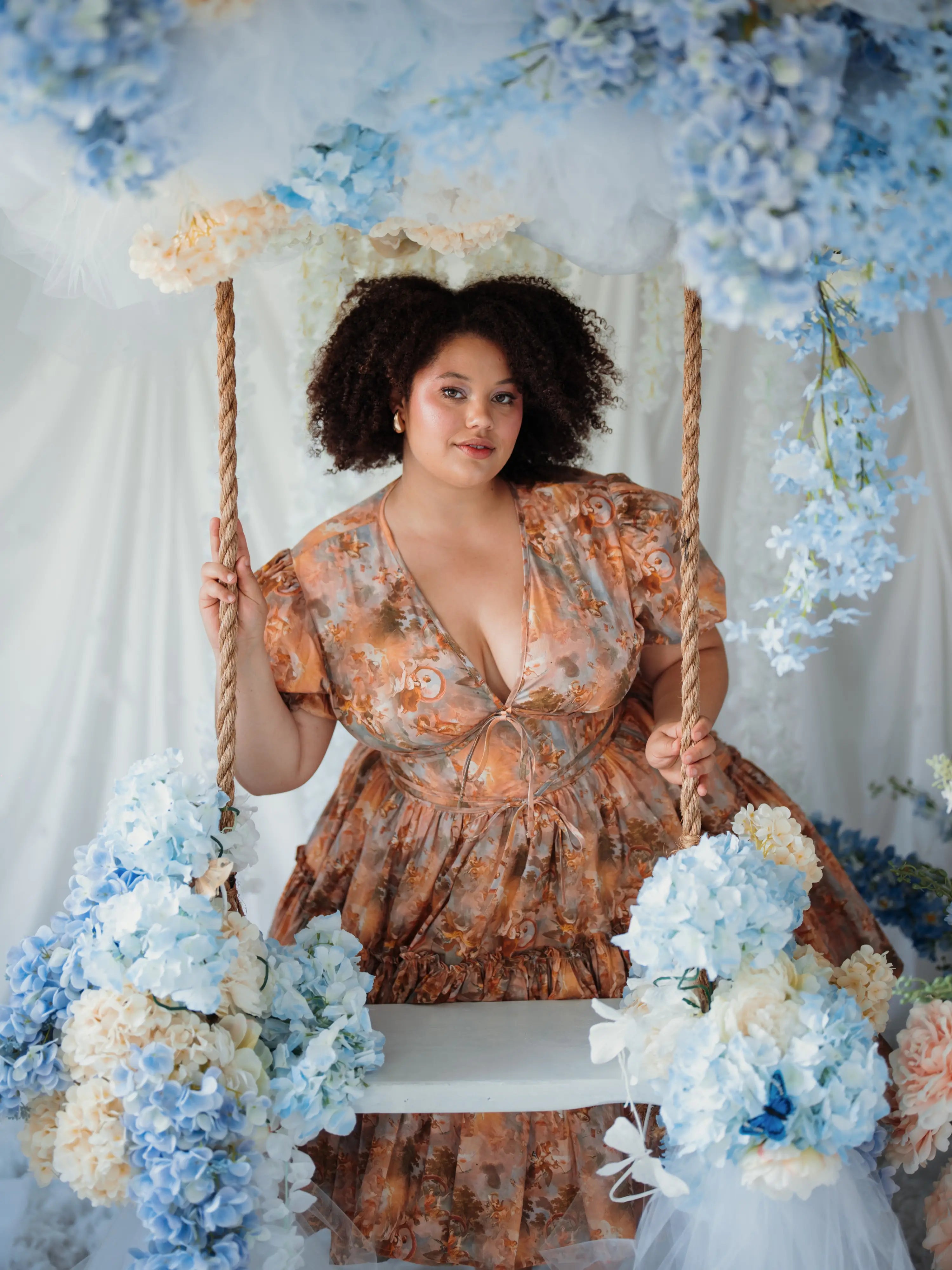 Woman in a floral dress sitting on a swing surrounded by blue and white flowers.