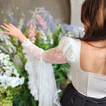 Woman in a white lace corset, The Guinevere Corset Top, featuring synthetic whale boning and bell sleeves surrounded by florals.