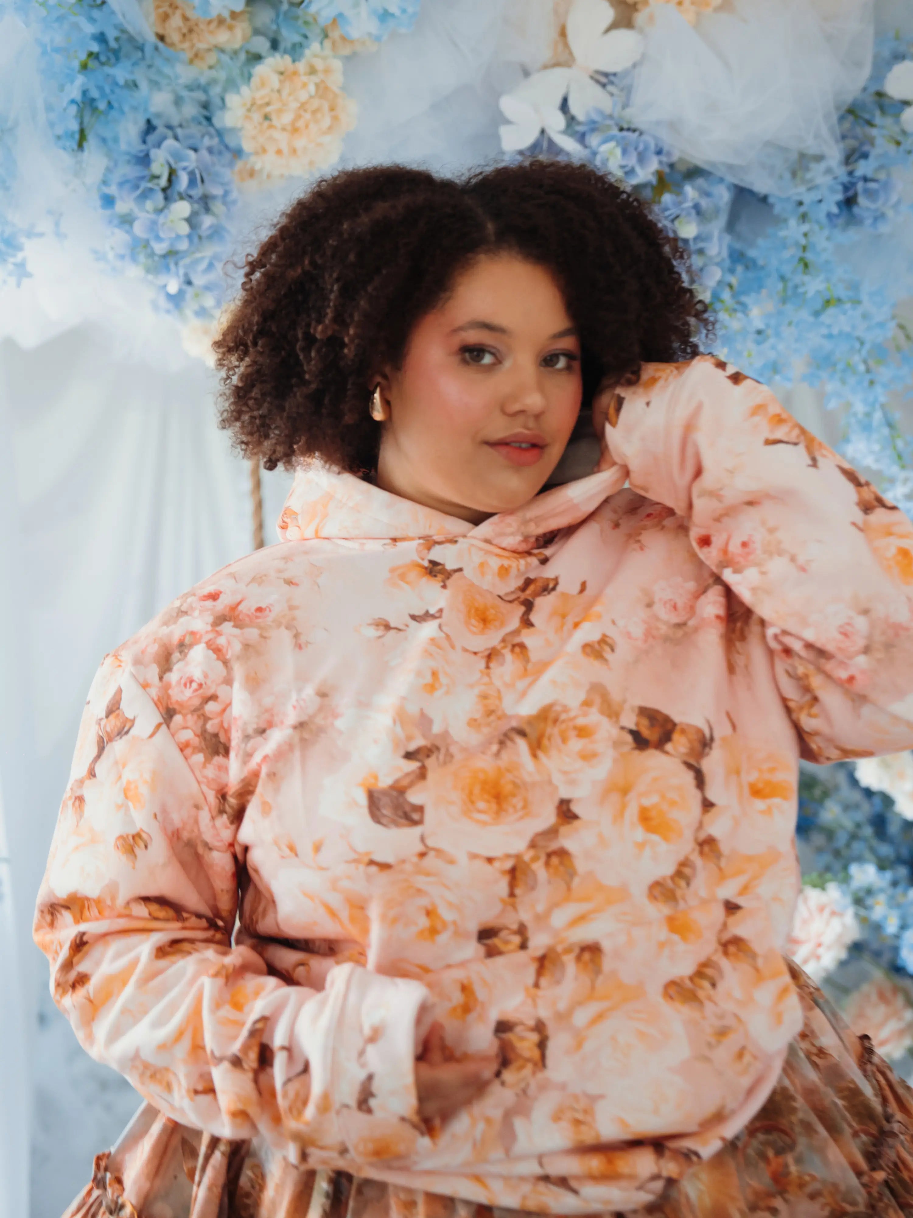 The Twilight Bloom Sleeper Hoodie in soft pink with peach floral print, modeled in a dreamy floral setting. Cozy oversized fit for romantic style lovers.