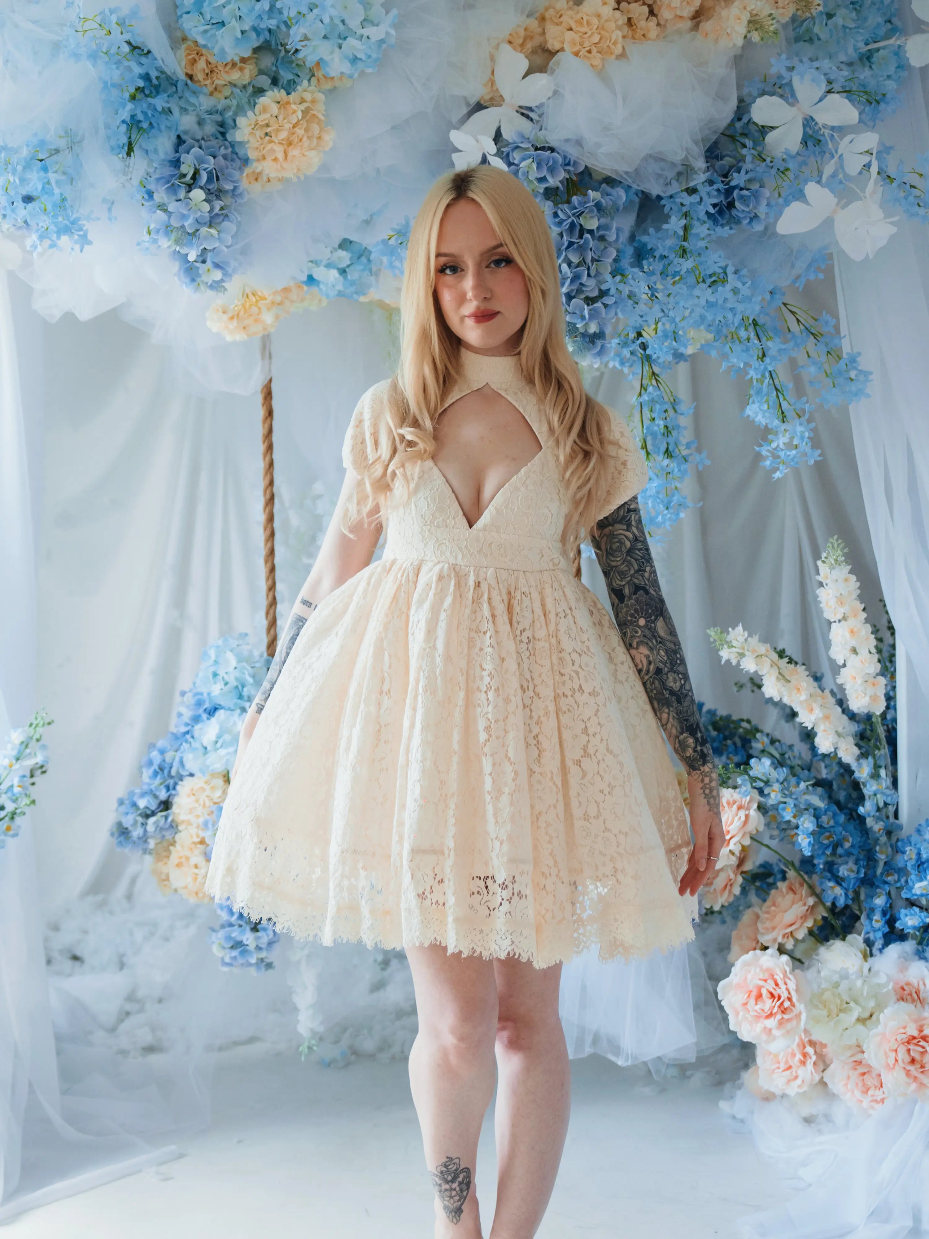 The Victorian Lace Tea Party Mini Dress in cream lace with puff sleeves and deep V-neck, styled in a floral setting. Women's Dresses category.