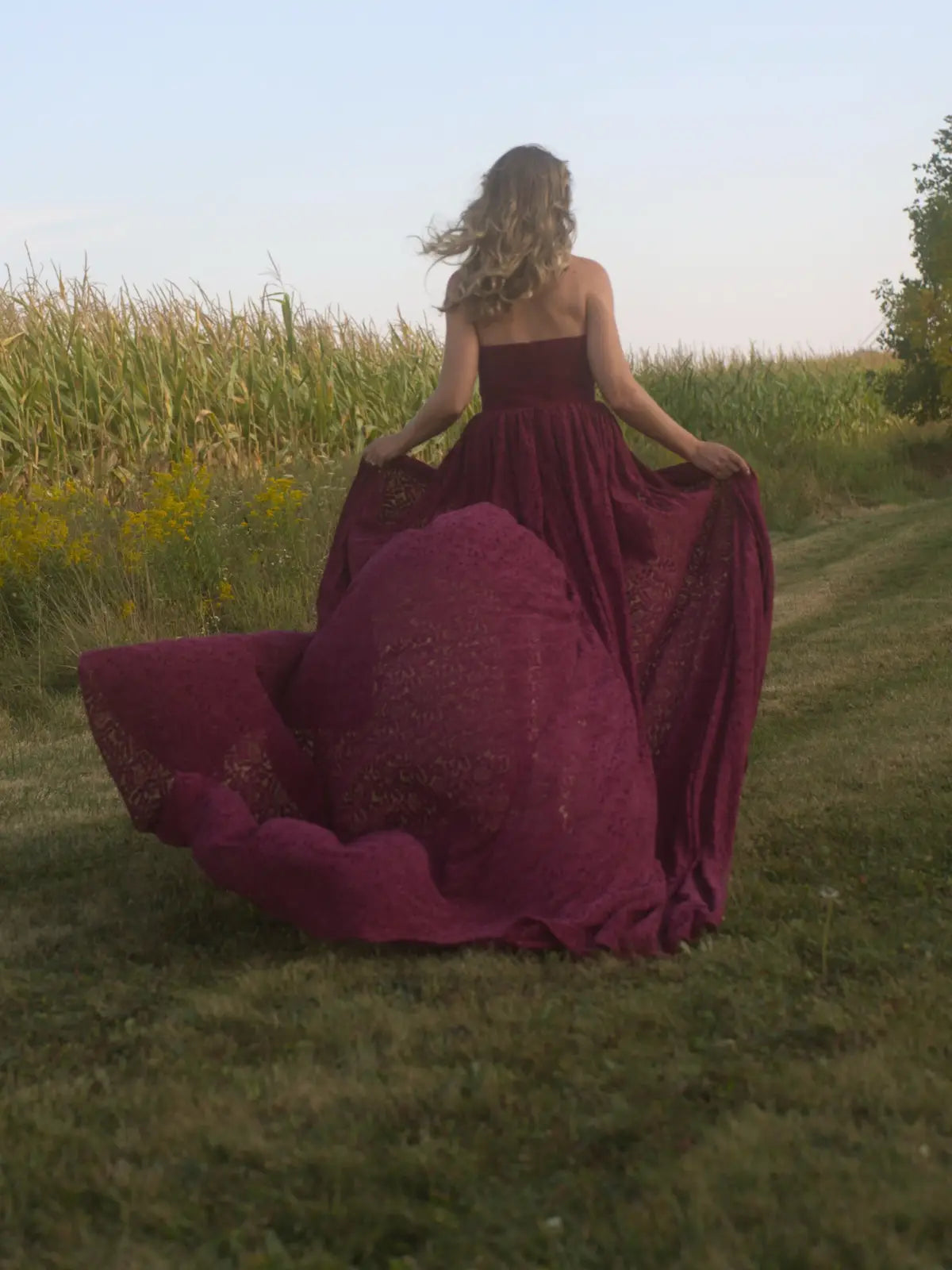 A woman in Ivory Sheep's Crimson Cathedral Gown running through a grassy field with trees in the background.