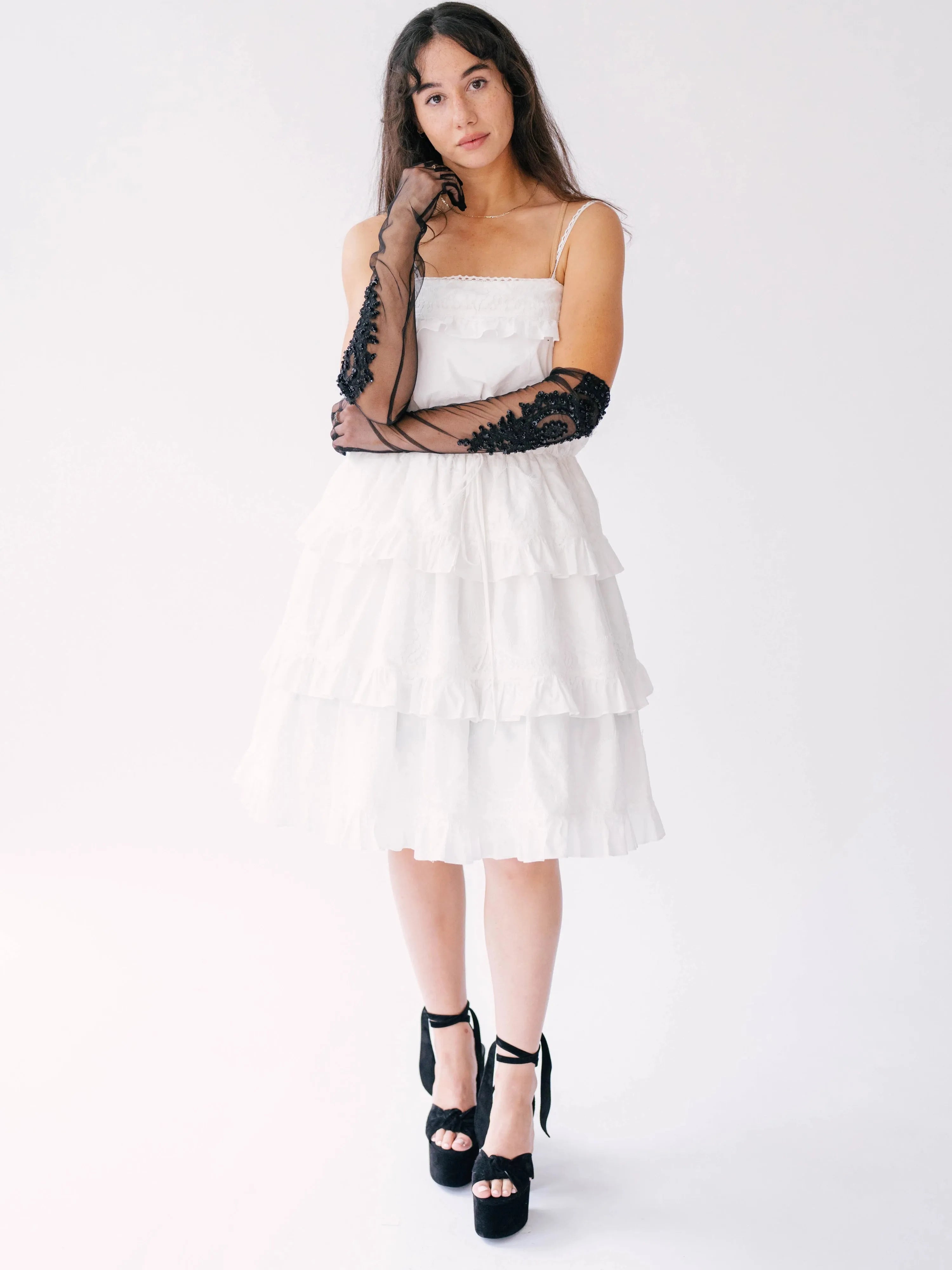 Woman wearing a white tiered sundress with spaghetti straps paired with black platform sandals and long black lace evening gloves.