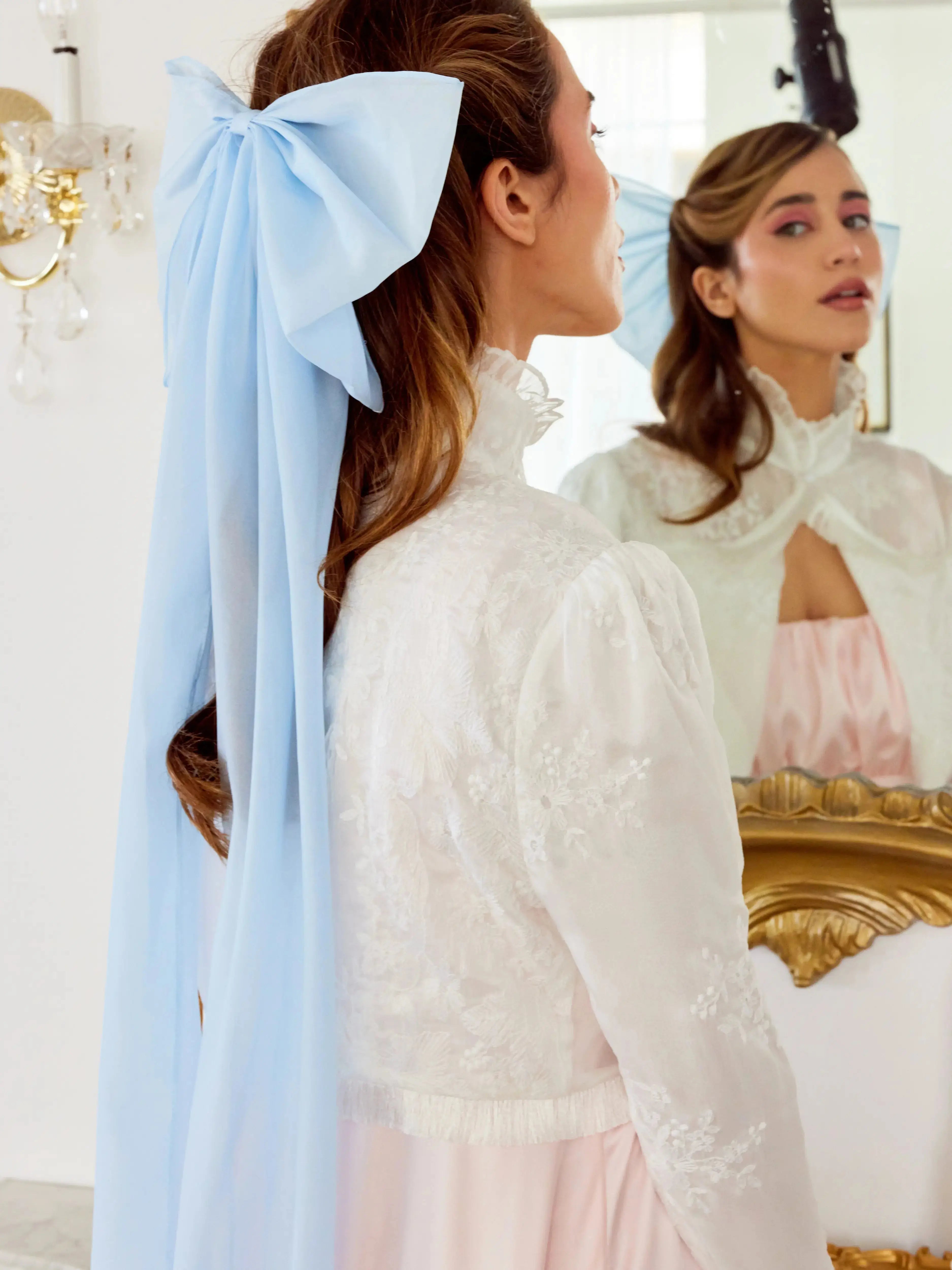 A woman with a large blue hair bow in her hair, wearing a white lace dress, standing in front of a mirror.