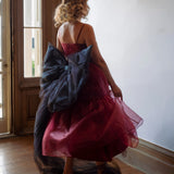 A woman in a red organza midi dress with a giant black tulle waist bow in a room with a wooden floor and white walls.