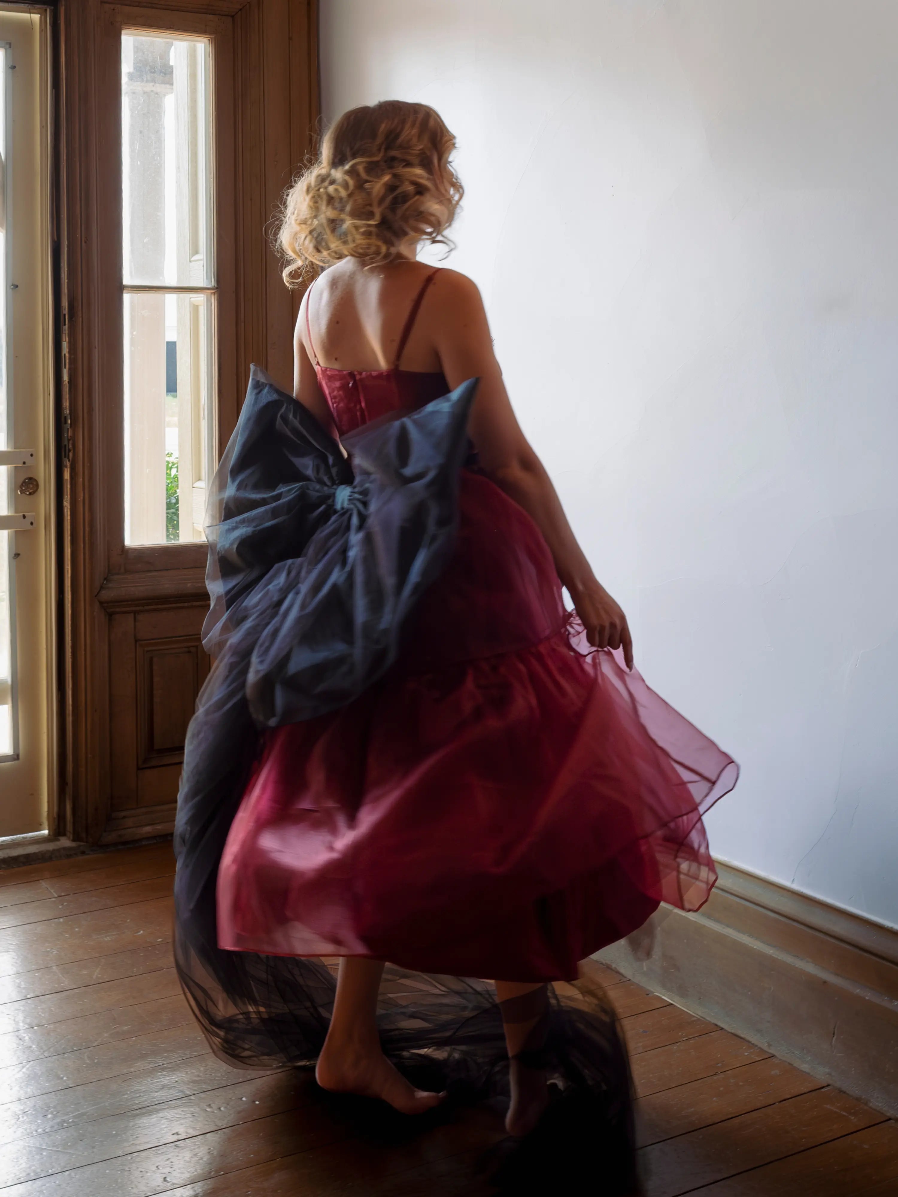 A woman in a red organza midi dress with a giant black tulle waist bow in a room with a wooden floor and white walls.