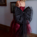 A woman wearing the Harlow organza midi dress with the black swan grand bow sash in an elegant room.
