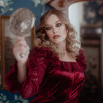 A woman in a red lace dress holding a hand mirror in a vintage room with floral wallpaper.