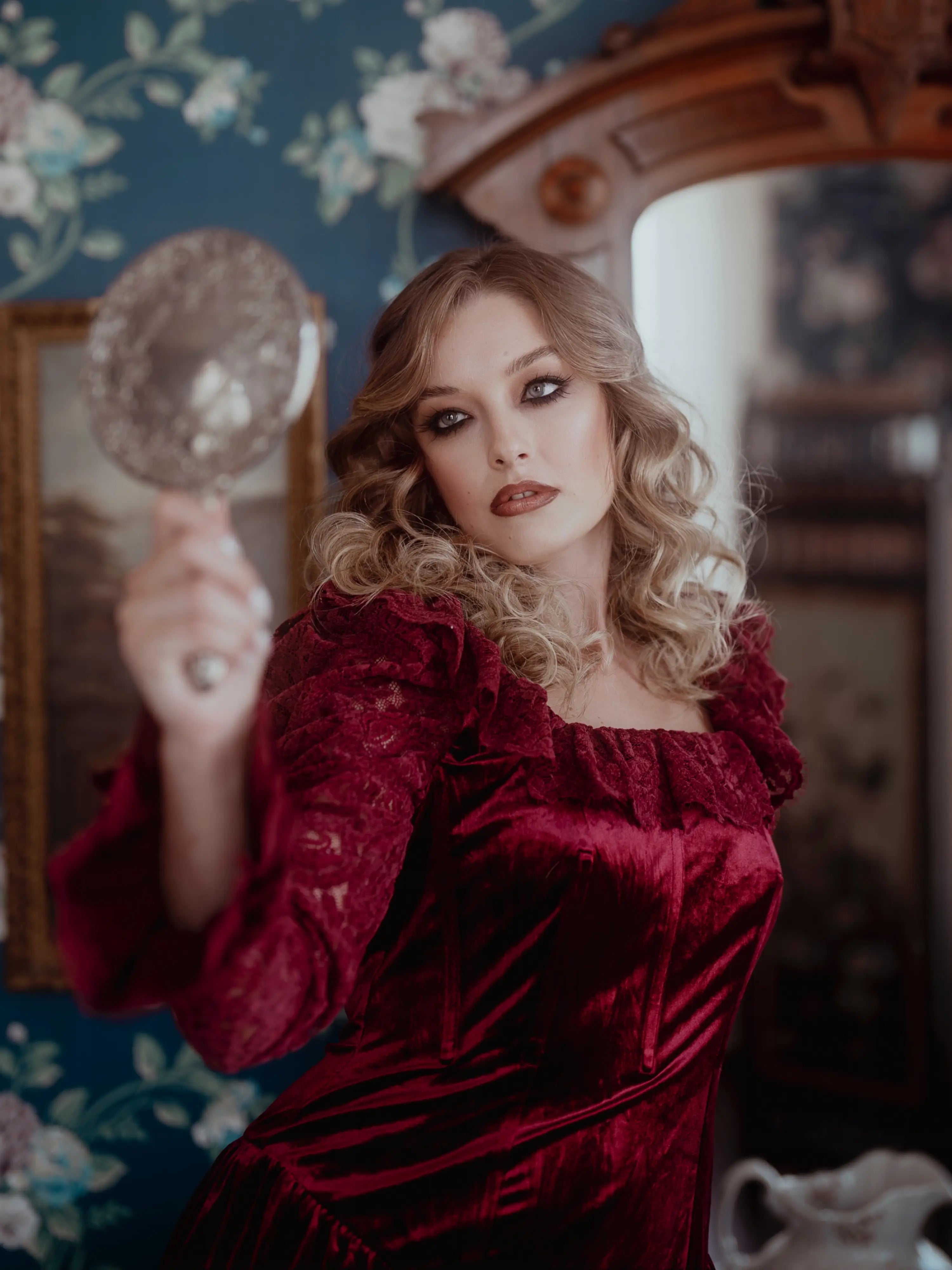 A woman in a red lace dress holding a hand mirror in a vintage room with floral wallpaper.