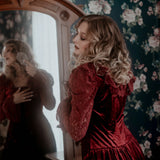 A woman in a red lace dress standing in front of a mirror with floral wallpaper.
