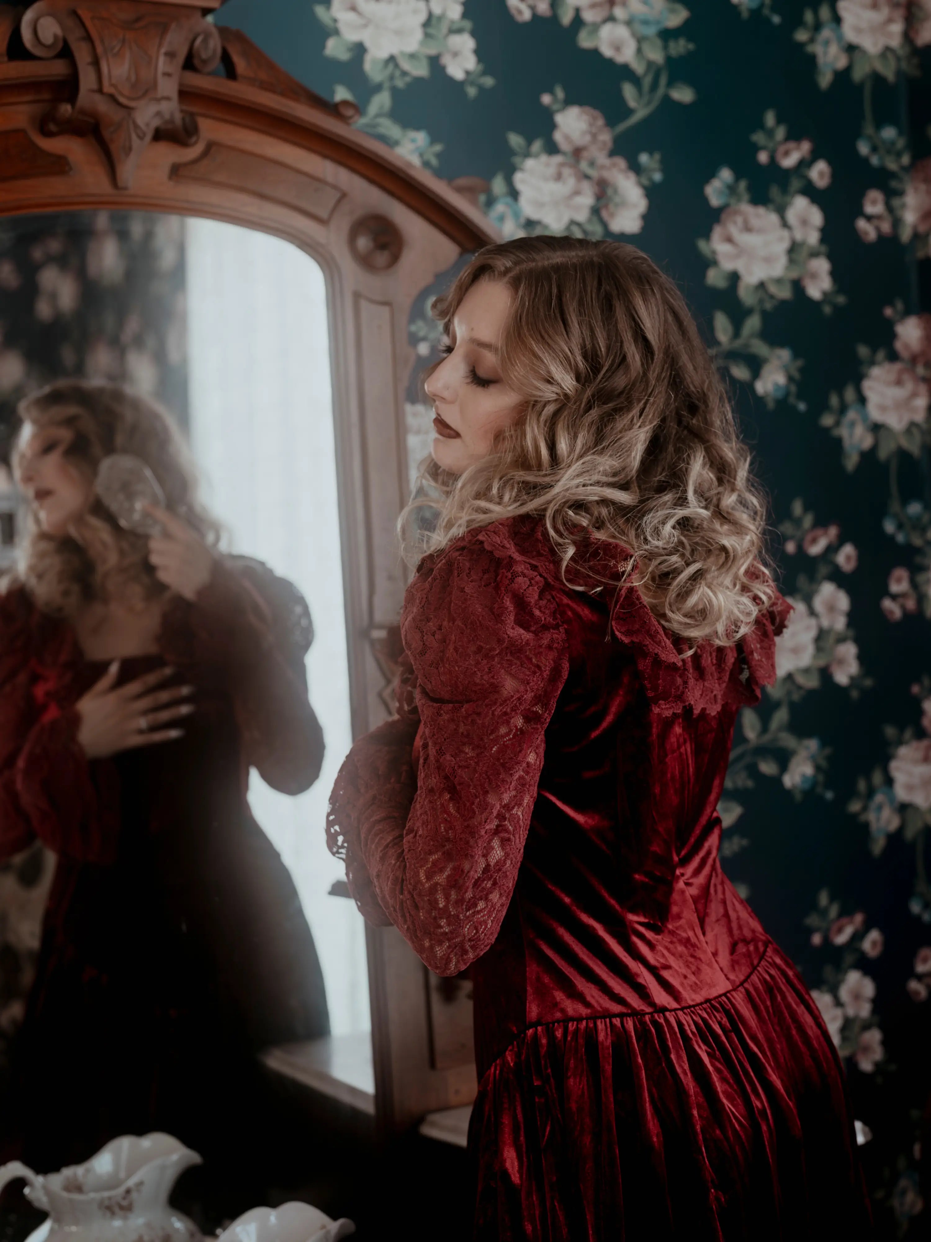 A woman in a red lace dress standing in front of a mirror with floral wallpaper.
