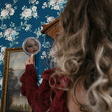 A woman holding a small mirror with floral wallpaper and a framed painting in the background.
