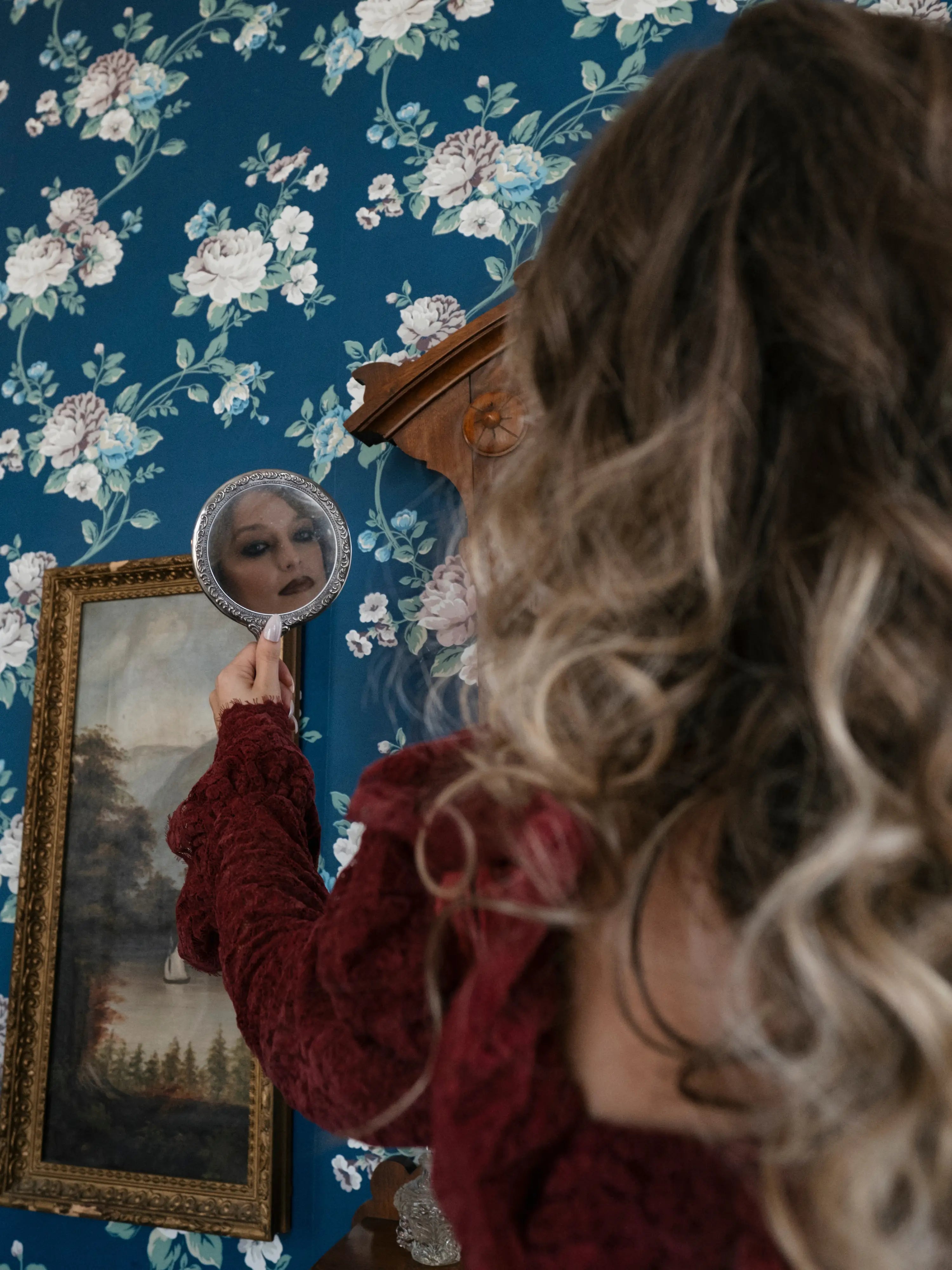 A woman holding a small mirror with floral wallpaper and a framed painting in the background.