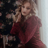A woman in a red velvet lace dress with floral wallpaper in the background.