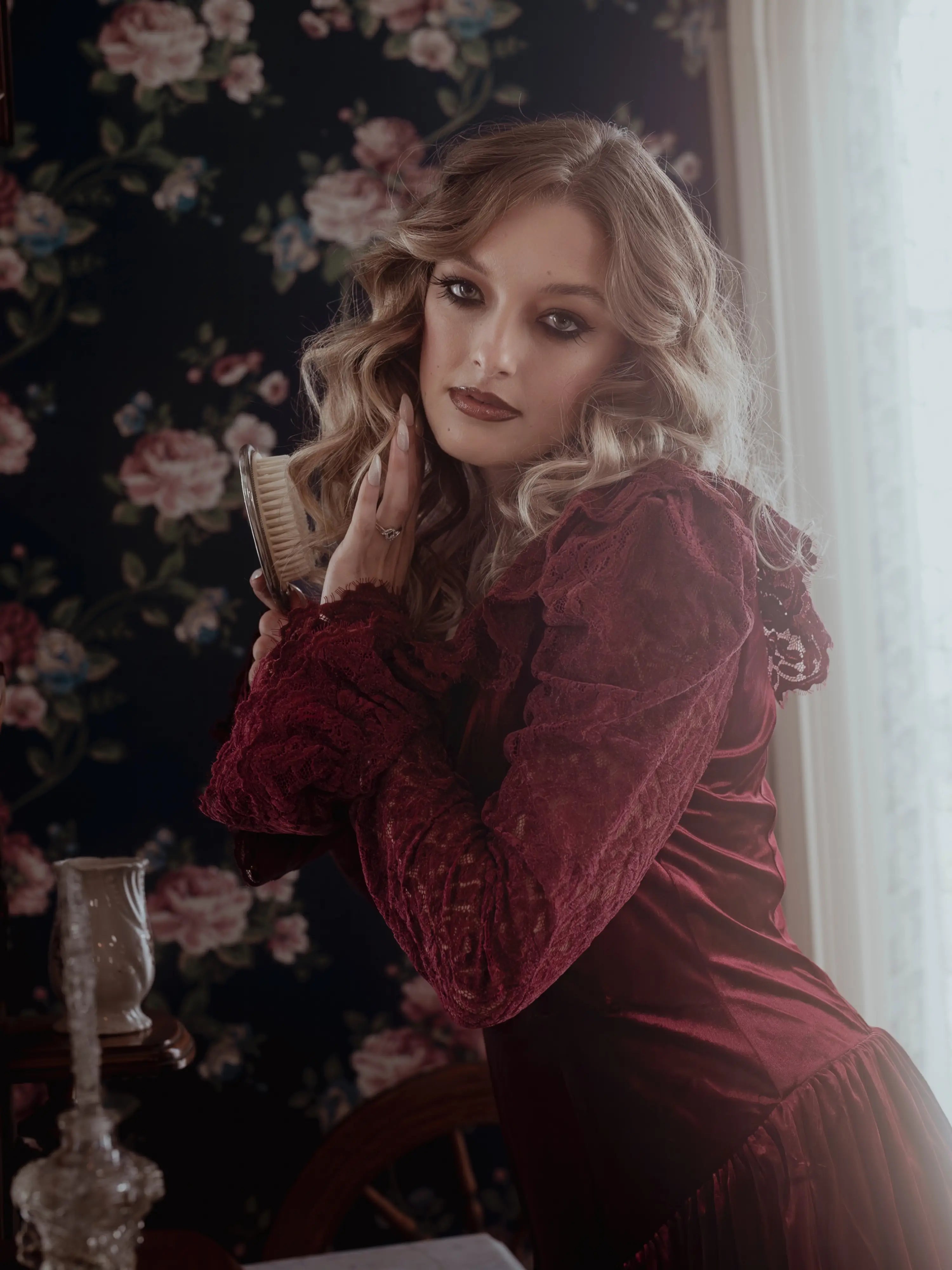 A woman in a red velvet lace dress with floral wallpaper in the background.