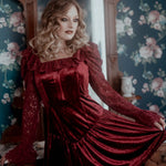 A woman in a red velvet lace dress standing in a room with floral wallpaper.