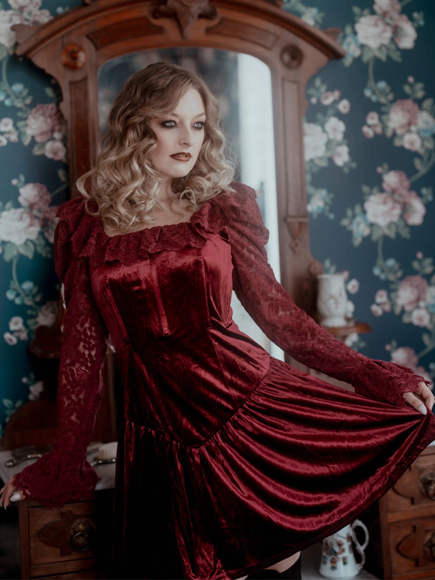 A woman in a red velvet lace dress standing in a room with floral wallpaper.