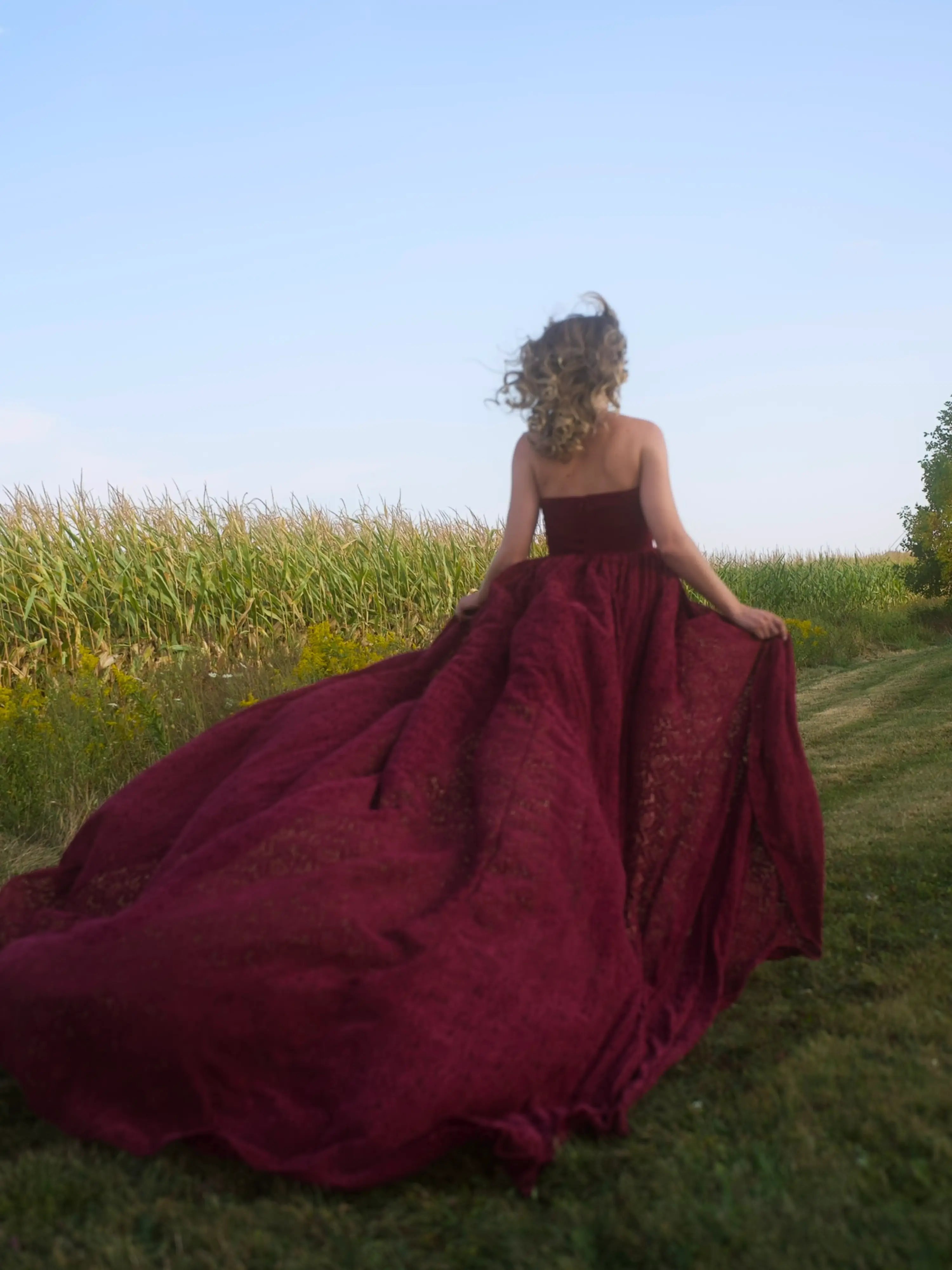A woman wearing the Cathedral Crimson Red Lace Dress is running in a field with a clear sky.