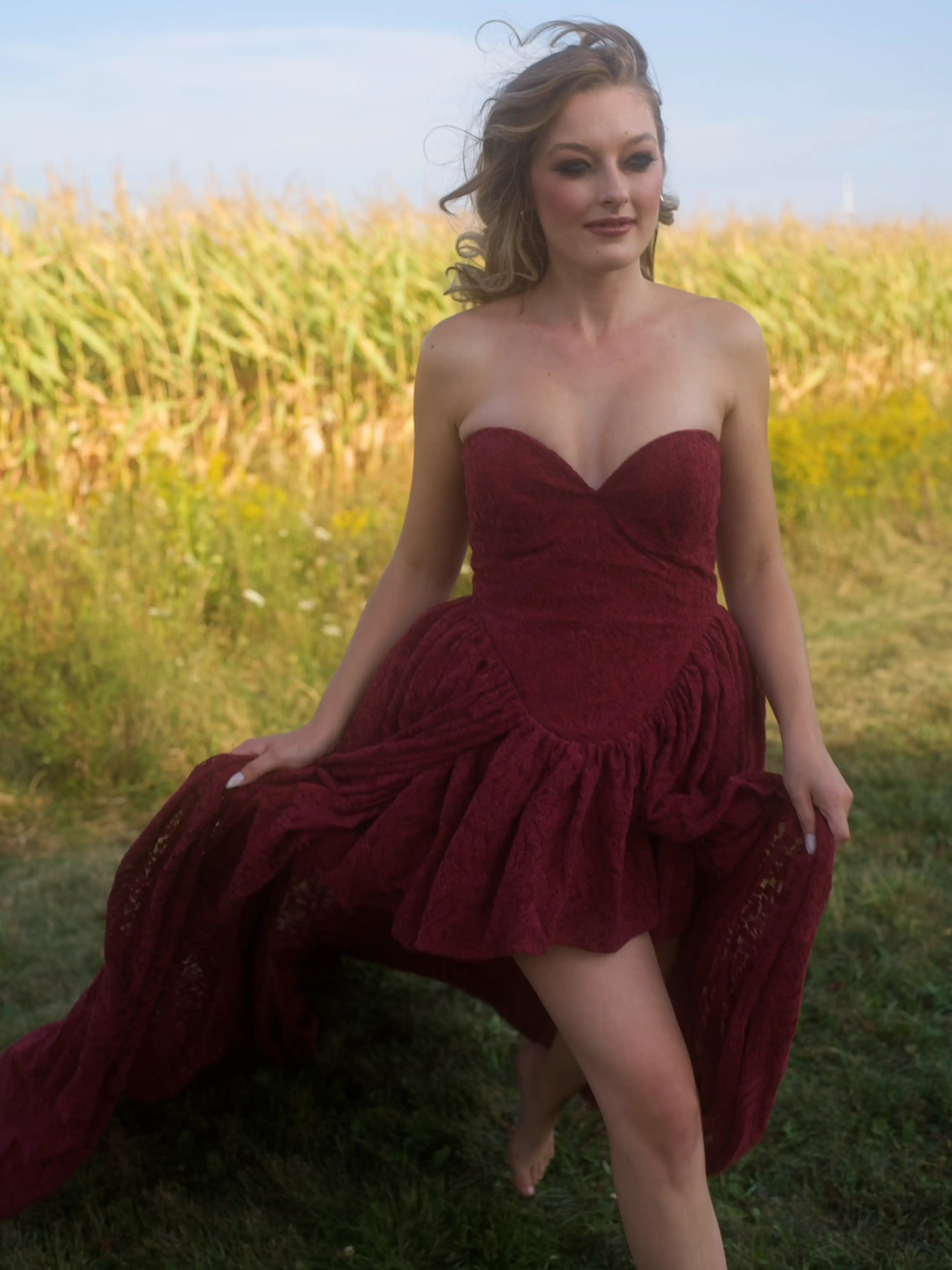 A woman wearing the Cathedral Crimson Red Lace Dress is walking in a grassy field.
