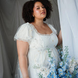Celestial Veil Dauphine Lace Gown with puff sleeves and embroidered stars, styled in a romantic setting with blue florals worn by a plus sized woman.