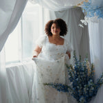 Plus sized woman in an embroidered lace gown holding a floral arrangement in a decorated room with white curtains and blue flowers.
