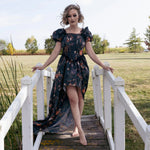 A woman in a dark autumn printed taffeta dress standing barefoot on a wooden bridge in a field