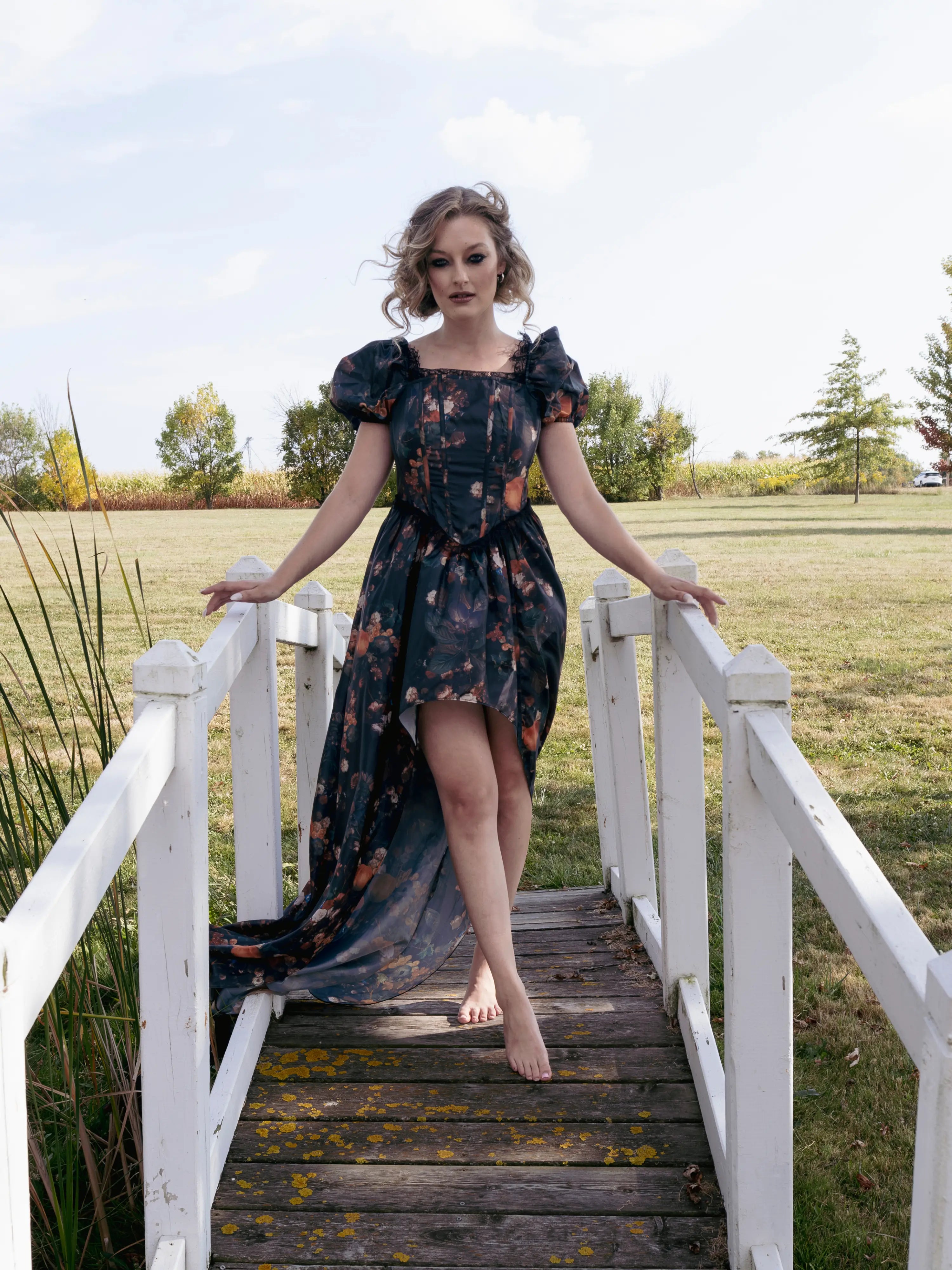 A woman in a dark autumn printed taffeta dress standing barefoot on a wooden bridge in a field