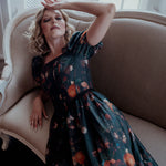 A woman wearing a still life printed taffeta dress sitting on an antique beige armchair with a window in the background