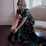 A woman wearing the still life Charlotte dress is sitting on the couch.