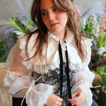 A woman in a ciela snow white shrug top featuring ornate lace trim and a black ribbon tie overtop a black corset mini dress.