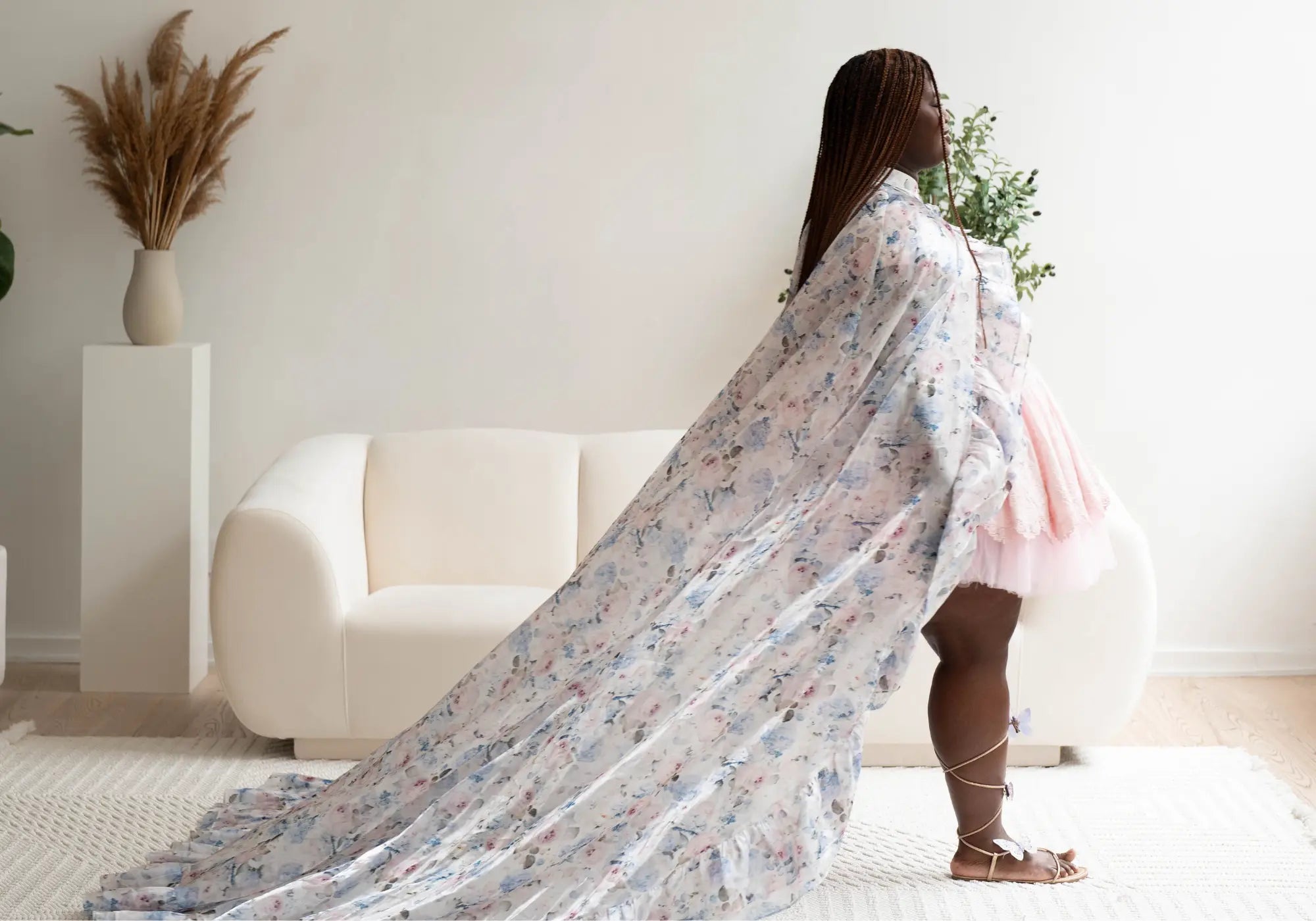 A woman is draped in a floral floor-length cape in a room with a white couch and decorative elements.