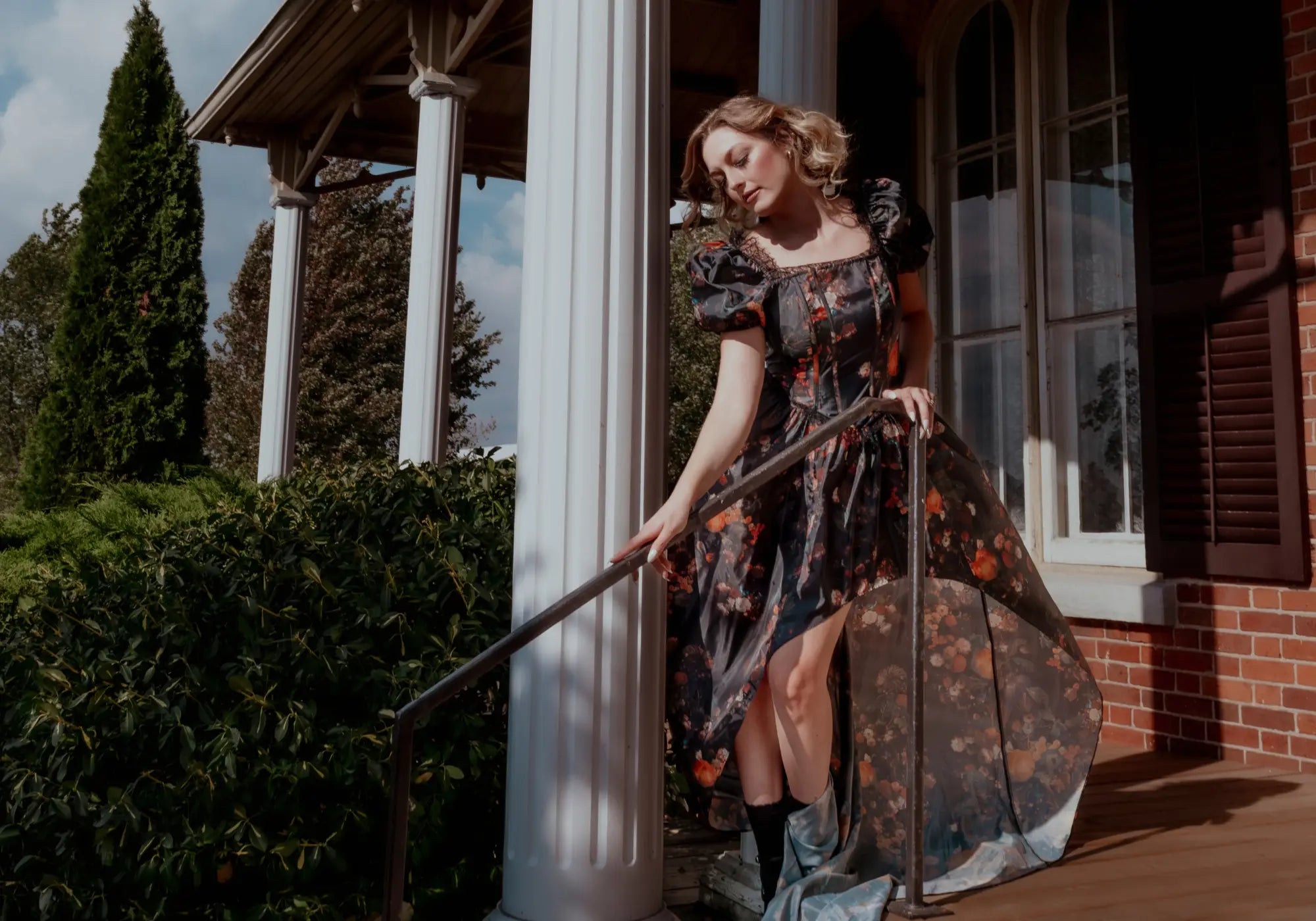 A woman in a floral cotton maxi dress holding a long step rail in front of a house with a porch.