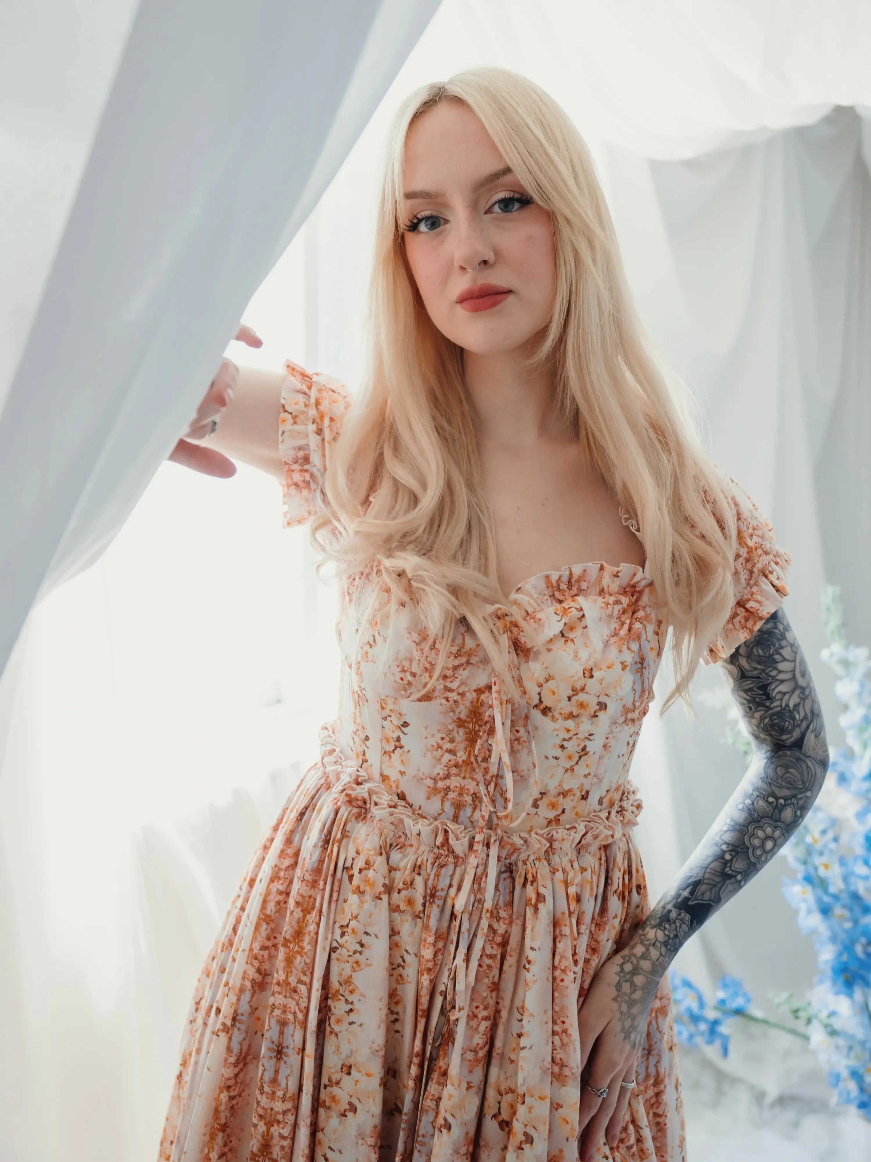 The twilight bloom meadow gown is a cotton gown worn by a model with her hand on her hip, showing ruffled bodice and romantic floral design in soft lighting.