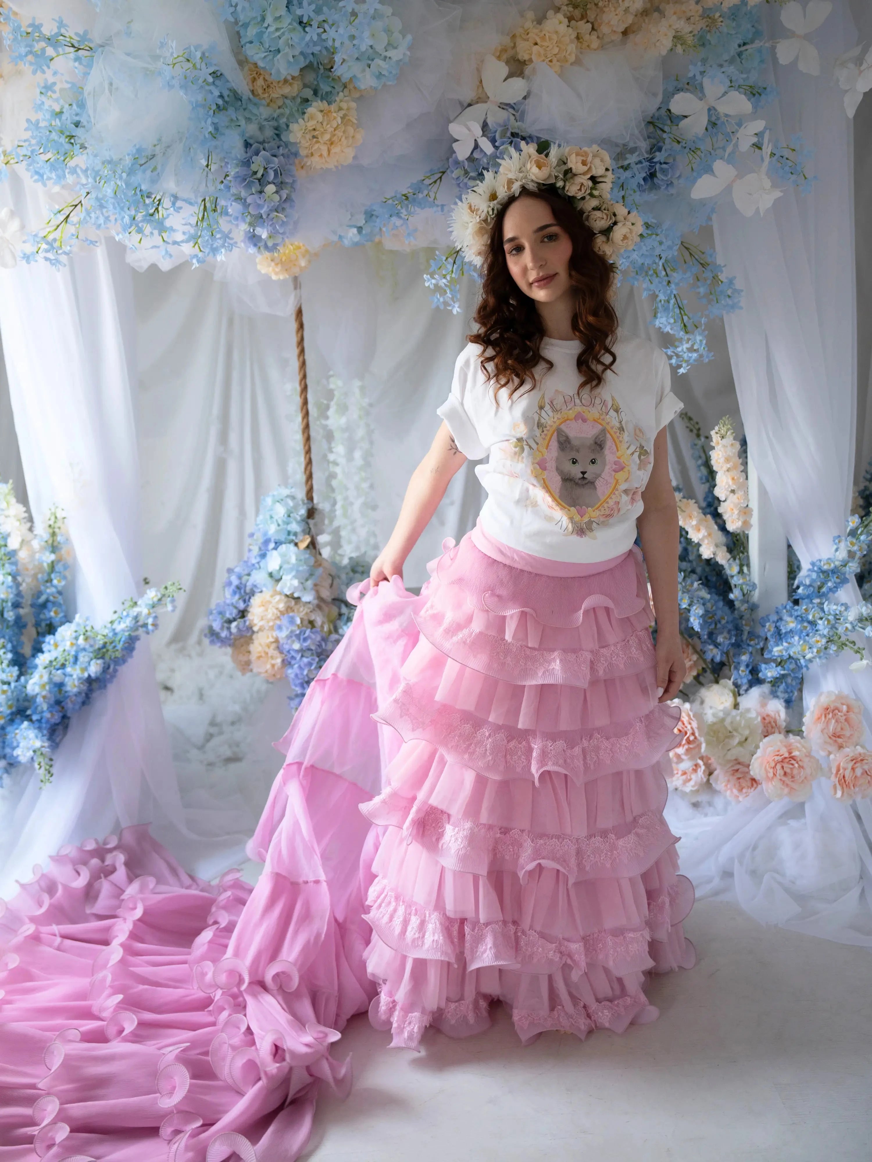 Front view of a woman wearing a t-shirt and a pink ruffle skirt in front of blue floral decorations.