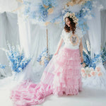 A woman in a white top with a kitten design and a pink ruffled skirt is standing in front of floral decorations.