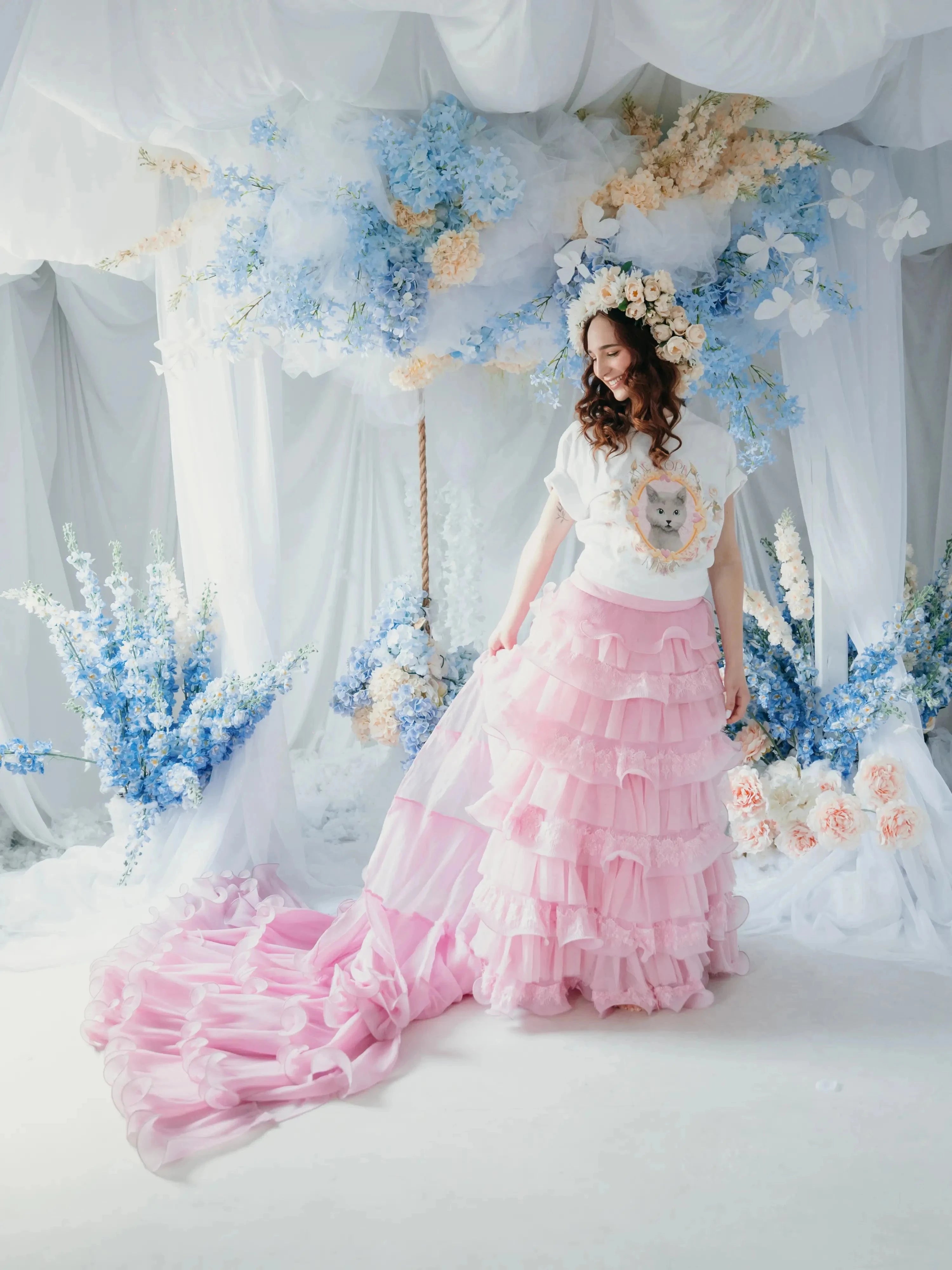 A woman in a white top with a kitten design and a pink ruffled skirt is standing in front of floral decorations.