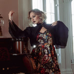 A woman wearing a stained glass satin mini dress and a black jacket, standing next to a piano in a room with large windows.