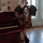 A woman wearing the Stained Rose Darling Dress, standing next to an old piano in a room with large windows.
