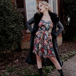 A woman wearing the Stained Rose Darling Dress and a black coat standing outdoors with a brick building and greenery in the background