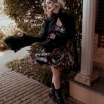 A woman wearing the Stained Rose Darling Dress and a black jacket, standing on a porch.