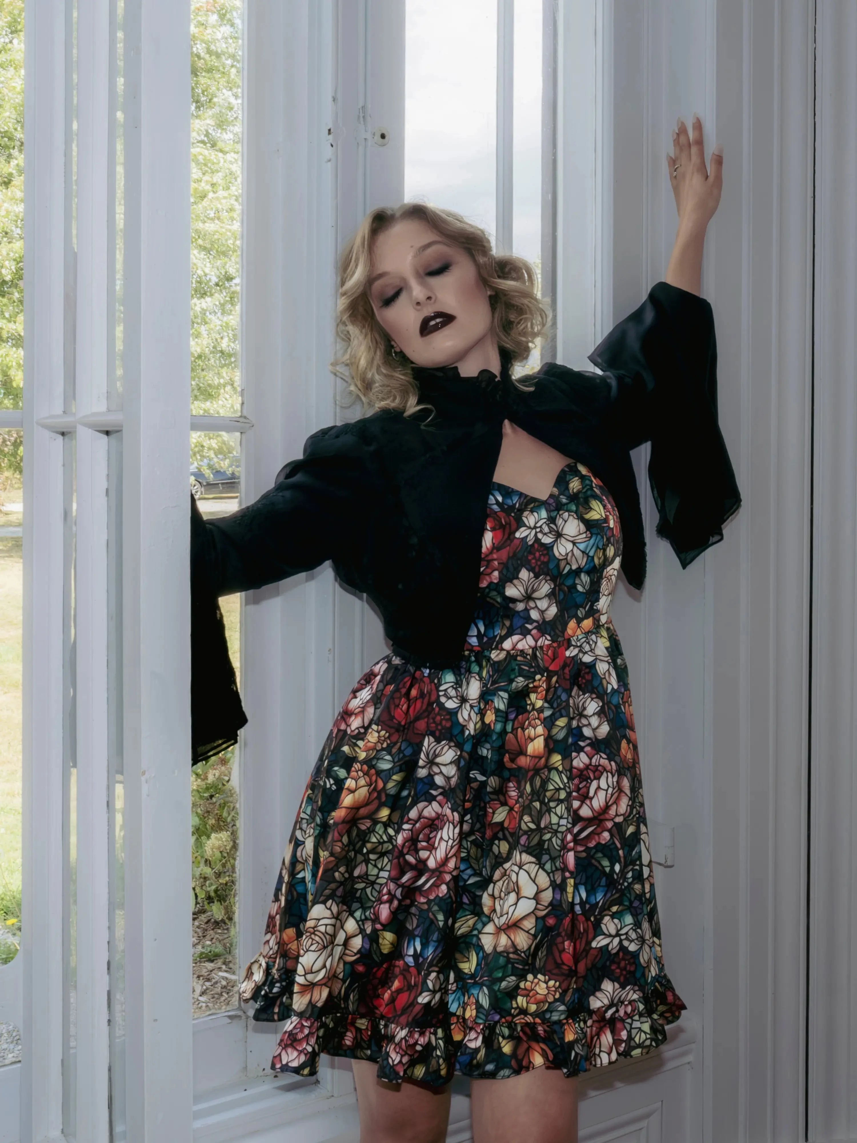 A woman wearing a stained glass satin mini dress and a black jacket stood before a white door.