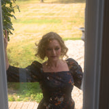 A woman in the still life dreamers cotton maxi dress standing in front of a glass door with a garden view.
