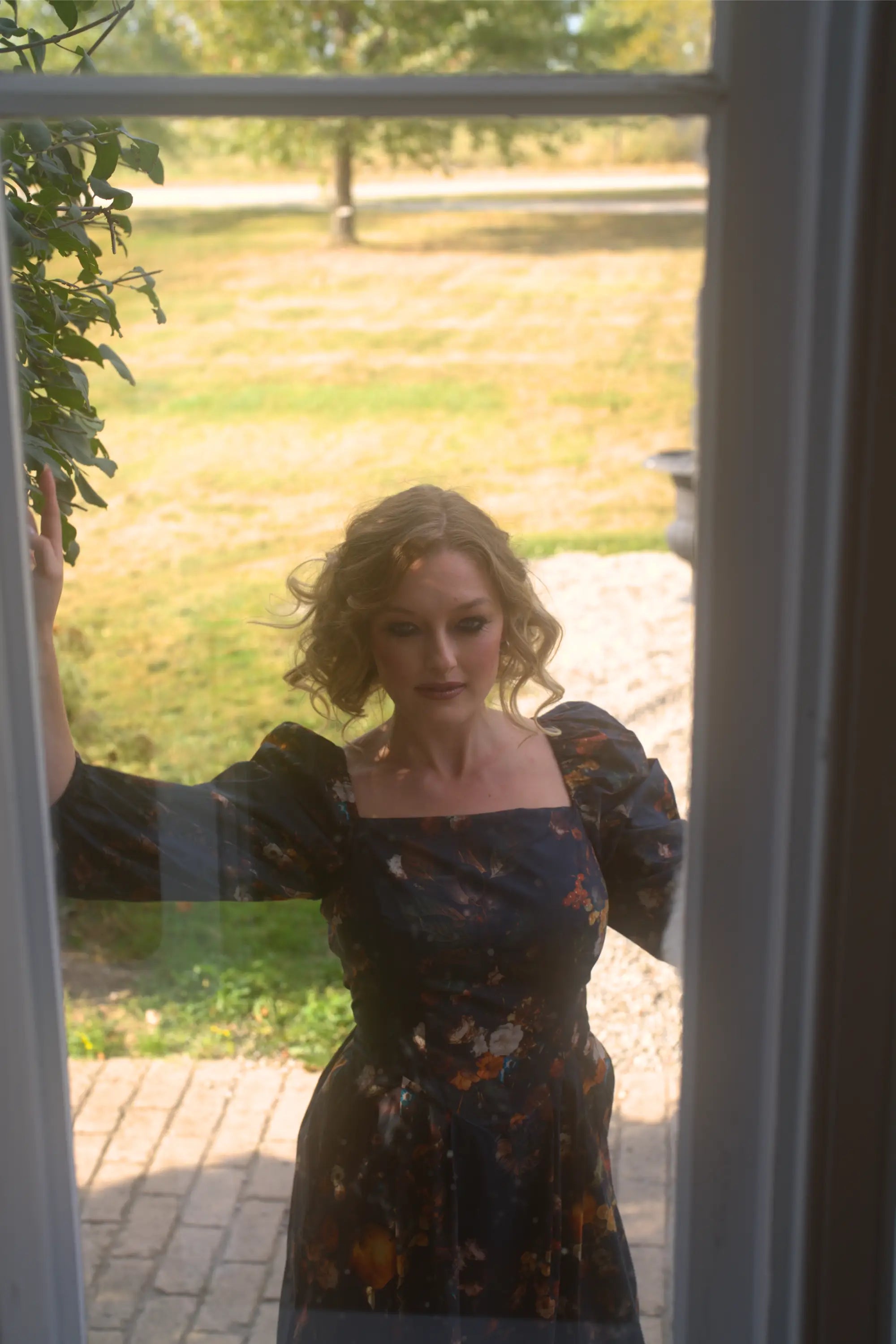 A woman in the still life dreamers cotton maxi dress standing in front of a glass door with a garden view.