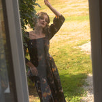 A woman in a floral cotton maxi dress standing outdoors, viewed through a glass door.