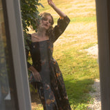 A woman in a floral cotton maxi dress standing outdoors, viewed through a glass door.